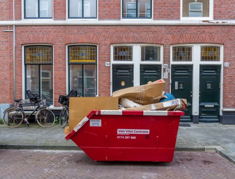 A large red waste skip with a white and red warning label on the side, positioned on a paved sidewalk in front of a row of three dark green front doors and tall, narrow windows with yellow caps, set within a brick building facade. The skip contains various discarded items, including flattened cardboard boxes, paper and plastic packaging, and miscellaneous rubbish. To the left of the skip, two bicycles are parked against the building's wall. The environment appears to be an urban residential street, with the building's brick exterior and the pavement visible beneath the skip. The scene is well-lit, with natural daylight illuminating the objects and surroundings. This image relates to rubbish removal services, which Waltham Forest House Clearance may provide in the local area around Waltham Forest, London, and nearby postcodes.