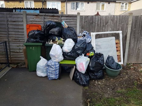 A collection of black, white, and patterned plastic rubbish bags containing household waste are piled on and around a green outdoor bench, which is located on a paved driveway in front of a wooden fence. Some bags are leaning against the fence and a green wheelie bin is positioned to the left side of the scene. Behind the rubbish, there is a white frame or panel and an unstacked wooden bed frame or piece of furniture. The background features the exterior of residential buildings with multiple windows and a tiled roof. The area appears to be in a suburban residential setting, with natural daylight illuminating the scene. This image visually documents waste ready for collection or disposal, relevant to rubbish removal services such as those offered by Waltham Forest House Clearance, serving Waltham Forest and surrounding areas.