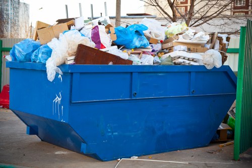 A large blue industrial skip bin filled with mixed rubbish materials, including crumpled white and transparent plastic and tissue paper, cardboard boxes, and some wooden pieces, is positioned outdoors on a paved area next to a green metal fence. The contents extend above the rim of the skip, which shows signs of minor wear and is set on a concrete surface. In the background, there are blurred residential buildings and bare trees, suggesting a typical urban area. The scene is lit by natural daylight, highlighting the various textures of the waste materials and the bright blue of the skip. This image visually represents a rubbish removal service, such as those offered by Waltham Forest House Clearance, aimed at clearing household or garden waste.