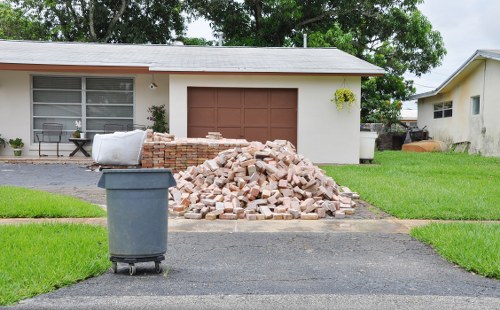 A pile of light brown brick debris and rubble is situated on the driveway in front of a single-story, cream-colored house with a brown garage door. The bricks are irregular in shape and appear partially broken or unused, forming a loose, uneven stack. To the left of the pile, a blue wheeled rubbish bin is positioned on dark asphalt, close to the curb. The house has a large rectangular window with white frames and curtains, and a small leafy plant growing in a window box near the right side of the house. The scene takes place in a residential area with lush green lawns on either side of the driveway, and other houses with similar single-story structures and light-colored exteriors visible in the background. The environment is well-lit, suggesting a clear day, and the presence of the debris indicates ongoing or recent rubbish removal or clearance activity by Waltham Forest House Clearance, specialists in local rubbish disposal services in Waltham Forest and nearby areas.