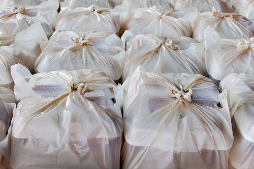A close-up view of numerous tightly tied white plastic bags filled with rubbish, arranged in rows on a flat surface, likely on a driveway or pavement in a residential or commercial area. The bags are made of semi-transparent plastic, revealing some of the contents inside, and are secured with beige twist ties or knots at the tops. The surface underneath appears smooth, possibly concrete or tarmac. In the background, additional bags are visible, with a glimpse of outdoor environment, indicating a waste clearance operation. The scene reflects a typical rubbish removal process, which Waltham Forest House Clearance services might manage, including collection and disposal of household or commercial waste in the local area.
